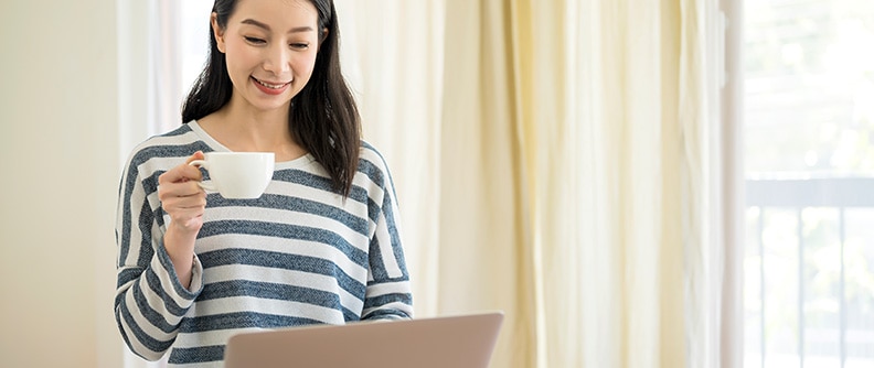 Mujer que sonríe y sostiene una taza mientras mira su computadora portátil