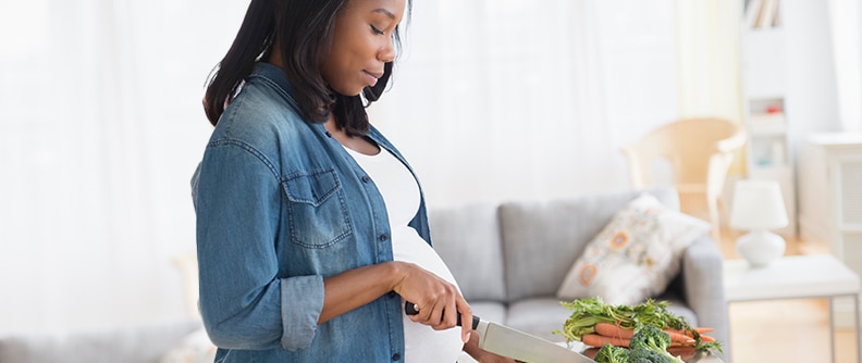Mujer de raza negra embarazada cortando verduras en la cocina.