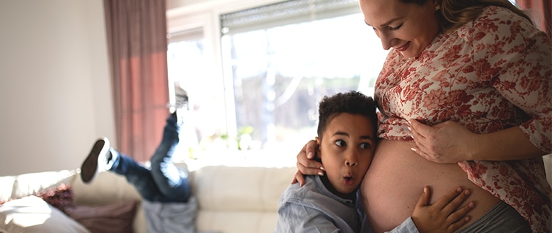 Niño sorprendido al escuchar los sonidos del vientre de su madre embarazada; otro hermano está saltando en el fondo de la imagen.