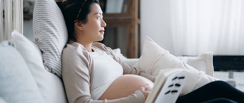 Mujer embarazada descansando con aire pensativo mientras lee un libro sobre bebés en el sofá de su hogar acogedor. A la espera de una nueva vida, el nacimiento y la crianza de los hijos.