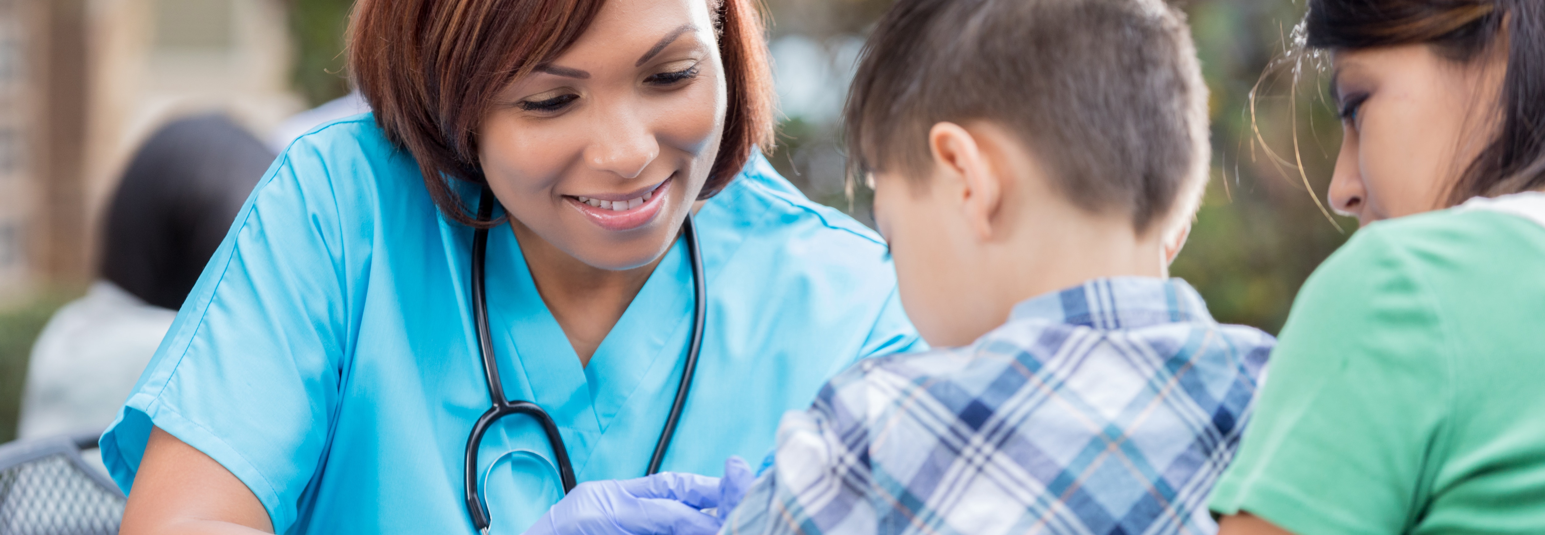 Enfermera afroamericana de mediana edad vacunando a un niño pequeño en una clínica gratuita al aire libre. El niño está sentado en el regazo de su mamá. La enfermera tiene puesto un uniforme médico y lleva un estetoscopio.