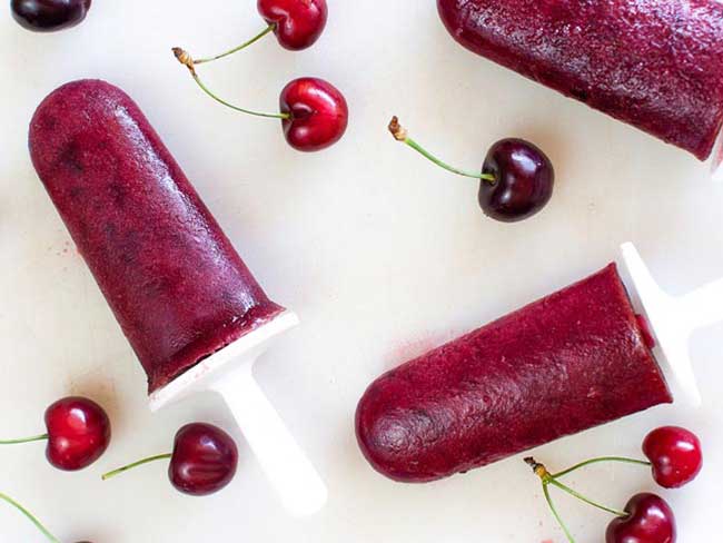 Fresh ripe cherries scattered around 3 homemade natural cherry popsicles.