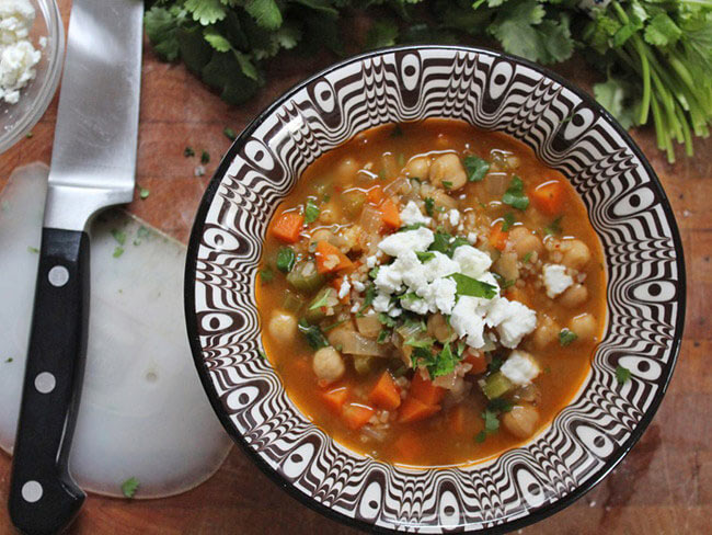 Bowl of spicy chickpea and bulgur soup on a wooden table with a knife and cutting board.