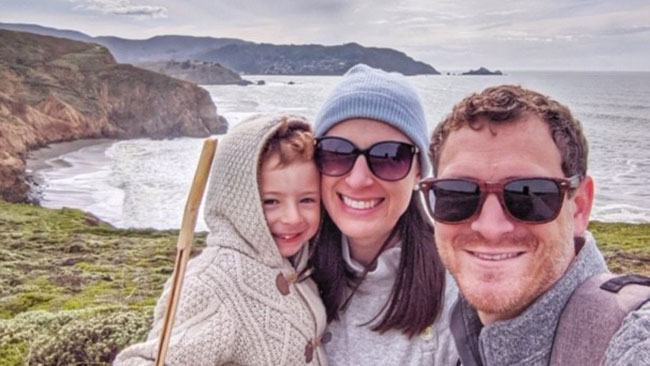 Two people standing together smiling and holding a baby, against a backdrop of cliffs and water.