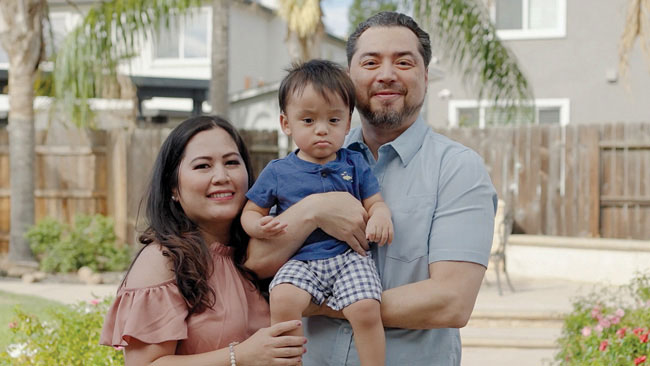 Parents holding their child and smiling in a backyard with greenery and a wooden fence. 