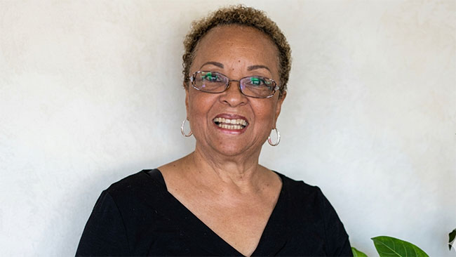 Persona con remera negra y anteojos sonriendo, con una pared blanca de fondo. 