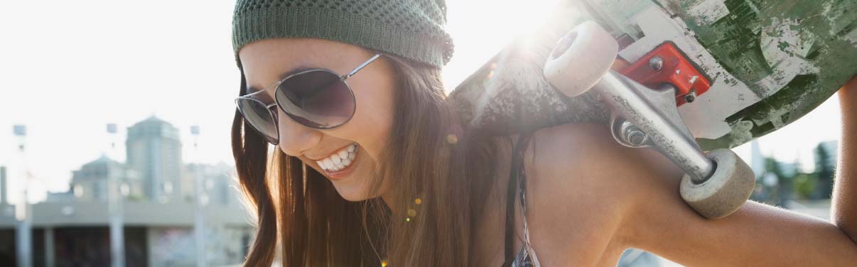 Una adolescente que sonríe y lleva puesto un gorro y unas gafas de sol, mientras sostiene una patineta