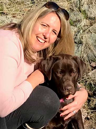 Mariann Stephens agachada abrazando a su labrador chocolate con una pared rocosa de fondo. 