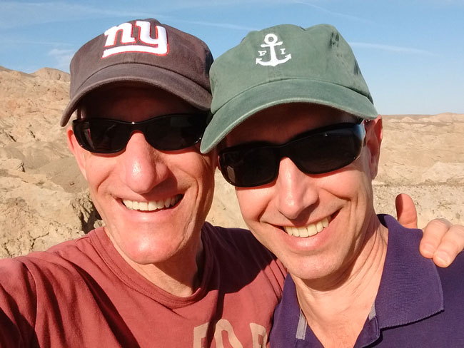 2 smiling men outdoors wearing sunglasses and caps.